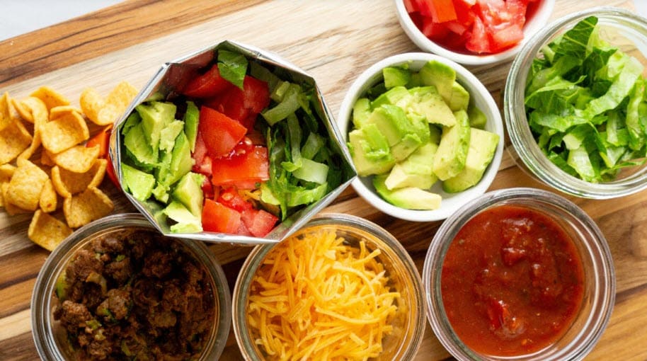 Ingredients for a taco or taco salad arranged in small bowls on a wooden surface, including corn chips, chopped tomatoes, diced avocado, shredded lettuce, seasoned ground beef, shredded cheese, and salsa.